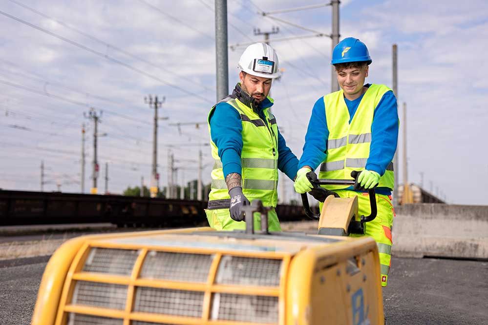 Ein Bauarbeiter erklärt einem Lehrling wie die Rüttelplatte funktioniert
