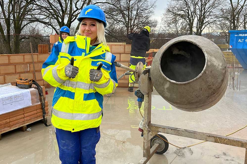 Ein Lehrling steht in Arbeitskleidung und mit Bauhelm neben einer Mischmaschine auf einer Baustelle