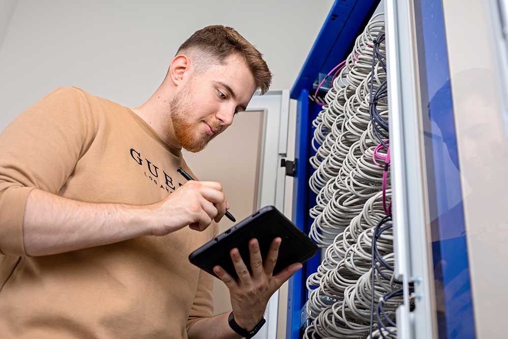 Ein Lehling steht mit einem Tablet vor einem Sicherrungskasten 