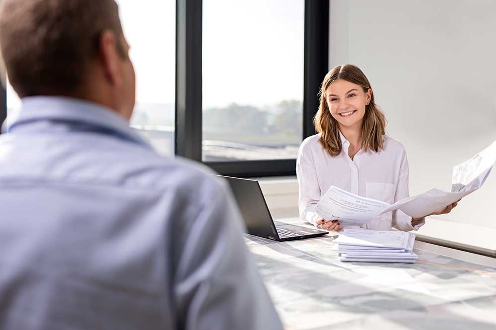Ein Lehrling mit Dokumenten in der Hand sieht lachend einen anderen Mitarbeiter an 