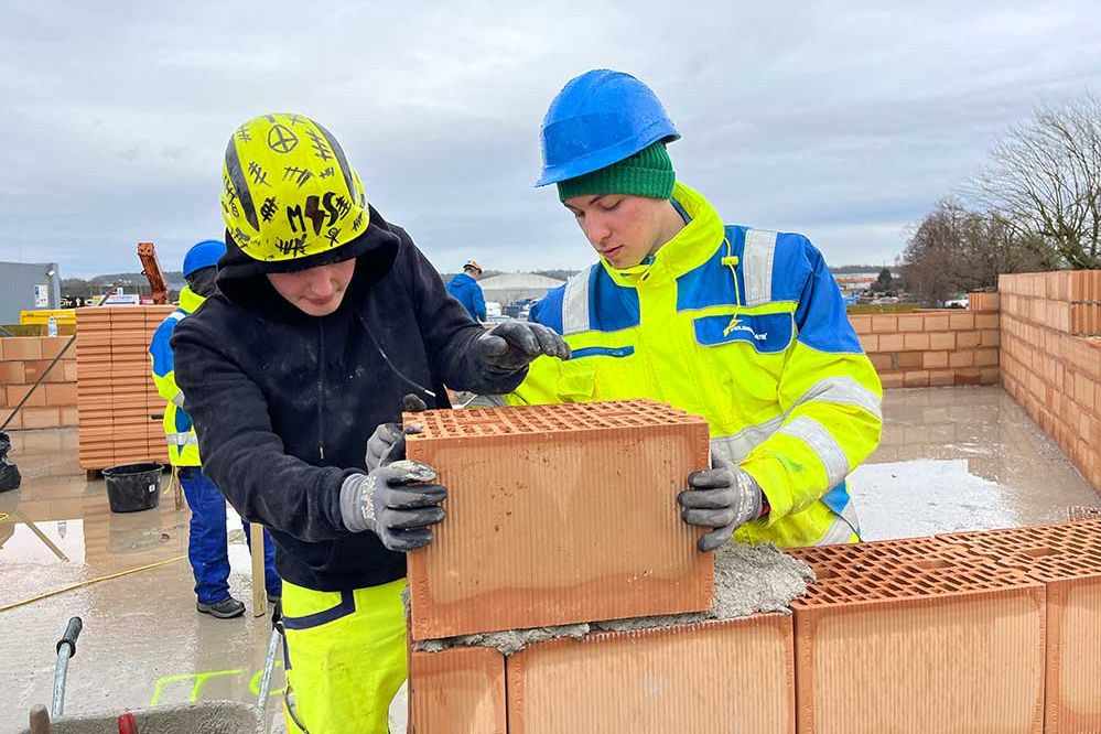 Lehre als Hochbauer: 2 Lehrlinge in Arbeitskleigung setzn auf einer Baustelle einen Ziegel auf eine Mauer