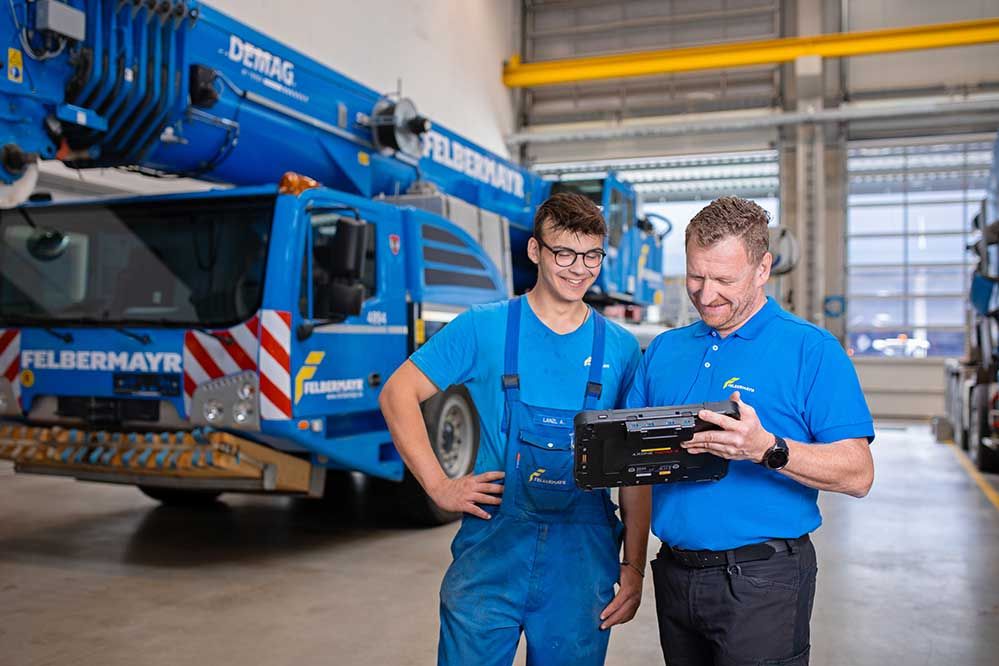 ein Mitarbeiter erklärt einem Lehrling etwas am Tablet in der Werkstatt. Im Hintergrund sieht man einen Mobilkran