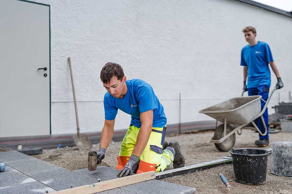 Lehre als Pflasterer: Ein Lehrling schiebt eine Schiebetruhe ein andereer Lehrling Pflastert den Boden