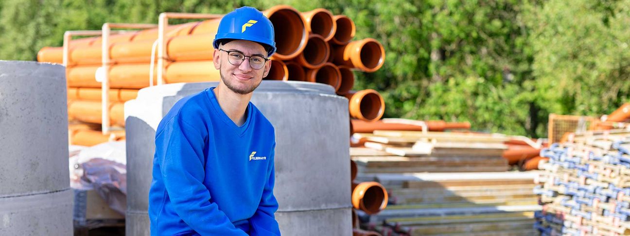 Lehre als Betonbauer: Ein Lehrling sitzt mit Arbeitskleidung und einem Bauhelm auf der Baustelle und lächelt in die Kamera
