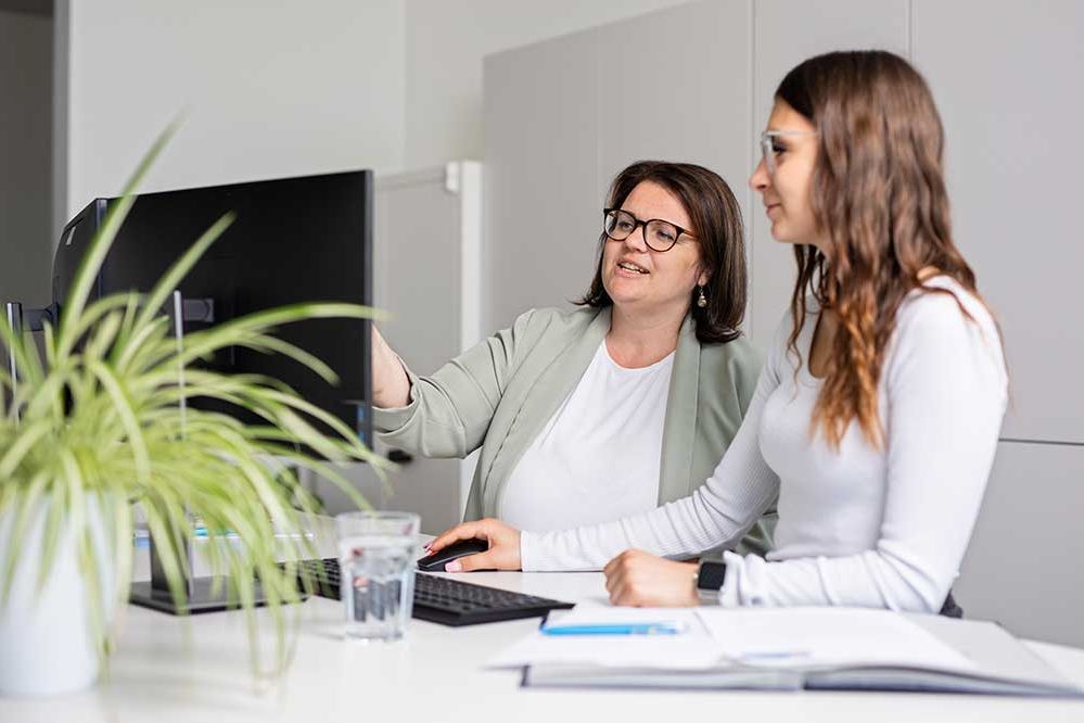 Eine Mitarbeiterin erklärt einem Lehrling etwas am Computer 