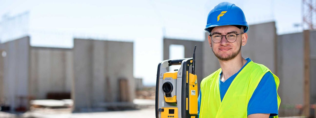 Lehre als Vermessungstechniker: Ein Lehrling mit Bauhelm auf einer Baustelle mit Vermessungsgerät