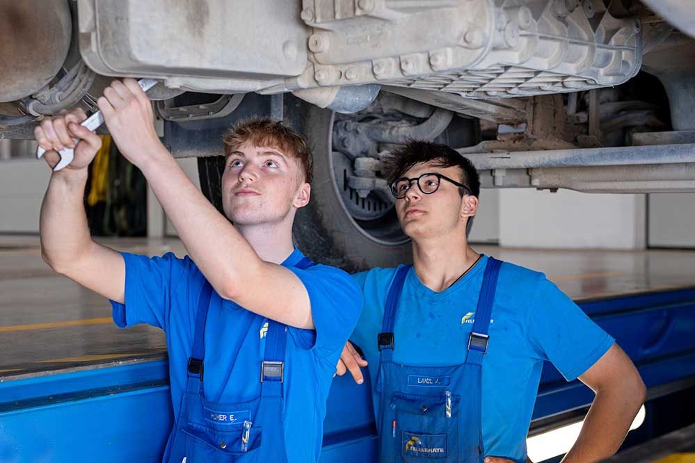 2 Lehrlinge stehn in der Montagegrube und schrauben an einem LKW