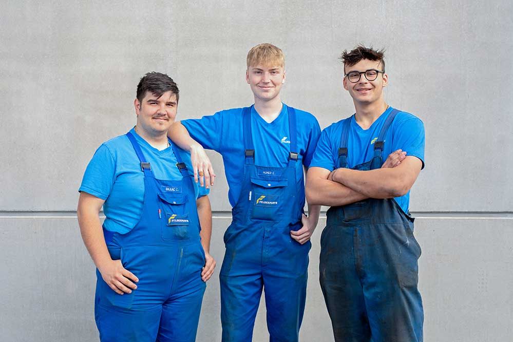 3 Lehrlinge stehen in Arbeitskleidung vor einer Betonwand