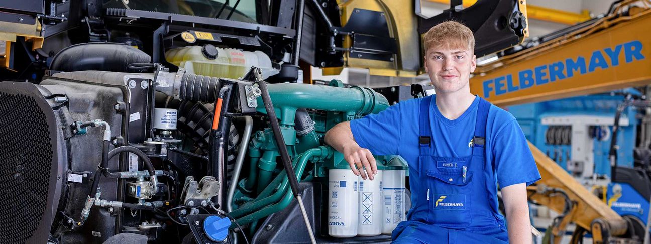 Lehre als Landmaschinentechniker und Baumaschinentechniker: ein Lehrling in Arbeitskleidung sitzt vor der geöffneten Motorhaube eines Radladers 