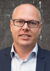 Ein Portraitfoto von dem Abteilungsleiter Diethard Schranz: Person in einem dunklen Sakko und einem hellen Hemd vor einer dunkel strukturierten Wand.