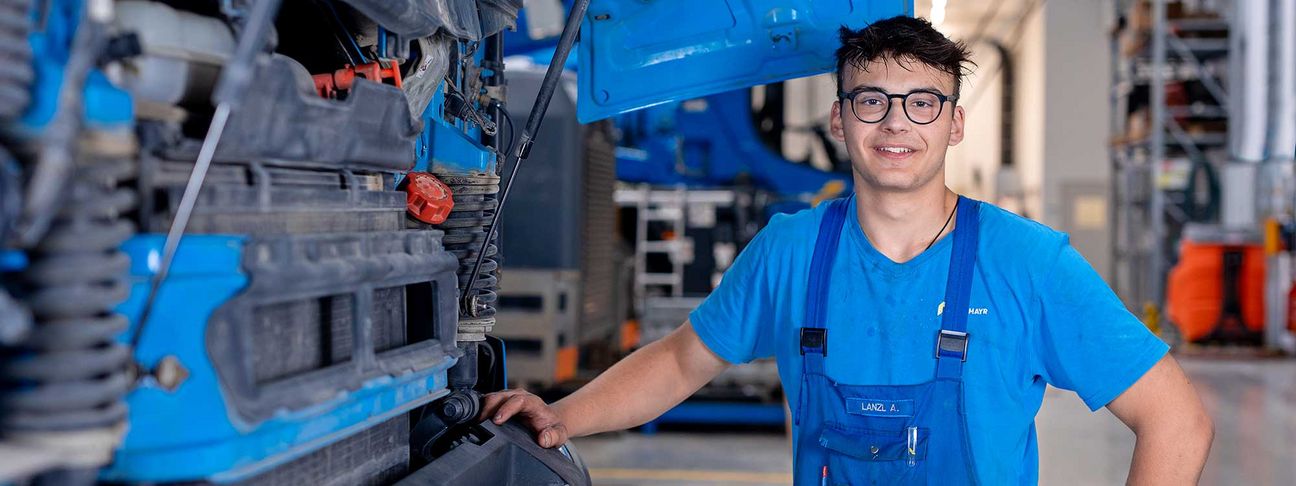 Lehre als KFZ Techniker: ein Lehrling in Arbeitskleidung steht vor der geöffneten Motorhaube eines LKW's 