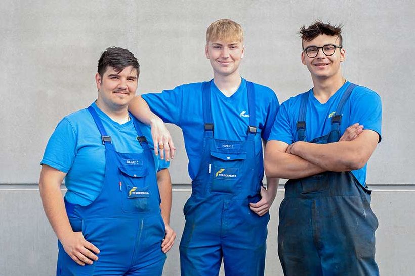 3 Lehrlinge stehen in Arbeitskleidung vor einer Betonwand