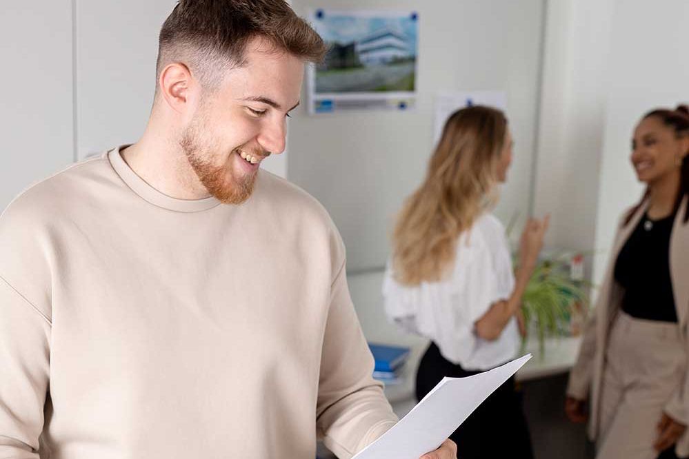 Ein Lehrling sieht sich lachend ein Dokument an, während sich im Hintergrund 2 Personen Unterhalten
