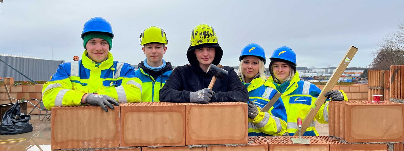 Lehre als Hochbauer: 5 Lehrlinge in Arbeitskleidung bei Regenwetter auf der Baustelle hinter einer gemauerten Wand 