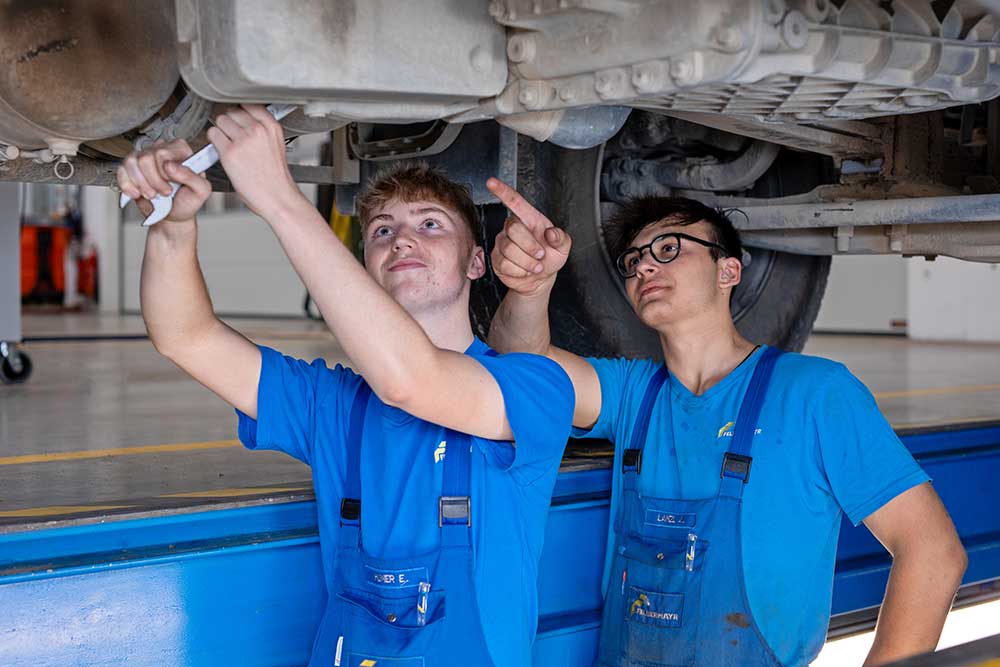2 Lehrlinge stehn in der Montagegrube und schrauben an einem LKW
