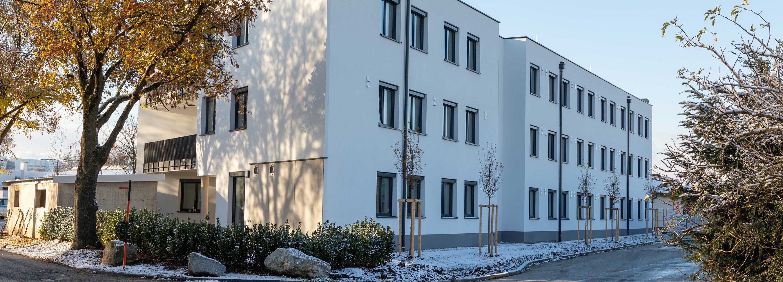 © Felbermayr/Weickinger Modernes, mehrstöckiges Gebäude mit weißen Fassaden und zahlreichen Fenstern an einer leicht verschneiten Straße, eingerahmt von Bäumen und Sträuchern.